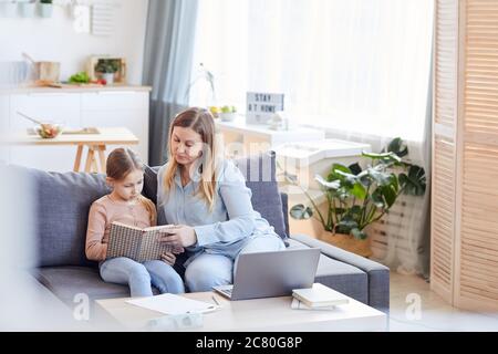 Weitwinkel-Porträt der liebevollen erwachsenen Mutter beobachten niedlichen Mädchen Buch lesen oder studieren, während auf der Couch sitzen in gemütlichen Innenraum, kopieren Raum Stockfoto