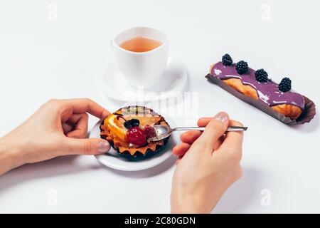 Nahaufnahme Frau isst Kuchen mit Löffel neben Tasse Tee und eclair auf weißem Tisch. Stockfoto