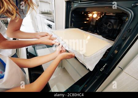 Nahaufnahme von Mutter und Tochter, die Backblech in heißen Ofen legen, wenn sie zu Hause Biskuitkuchen machen Stockfoto