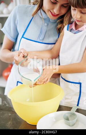 Lächelnde Mutter und Tochter Gießen Glas warme Milch in großen Plastick Schüssel, wenn Pfannkuchenteig Stockfoto
