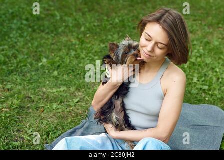 Ein Mädchen mit ihrem Hund sitzt im Park. Ein kleiner Yorkshire Terrier Hund ist in den Armen seiner Besitzerin und leckt ihr Gesicht. Küsse mit einem Hund. Stockfoto