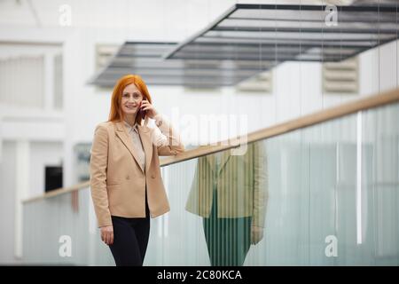 Porträt einer reifen rothaarigen Geschäftsfrau, die mit dem Smartphone spricht und die Kamera anschaut, während sie auf dem Balkon im weißen Bürogebäude steht, Stockfoto