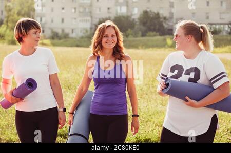 Drei glückliche junge Frauen tragen Yoga oder Turnmatten, wie sie über ein Feld in einem städtischen Park zu schlendern und zu plaudern und lächeln auf ihrem Weg zum Training in einem Stockfoto