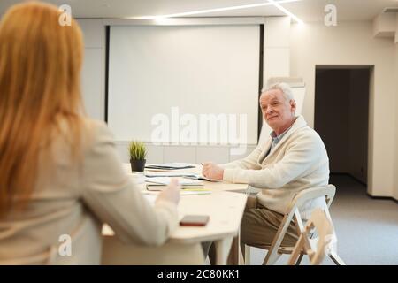 Porträt eines weißhaarigen Geschäftsmannes, der im Konferenzraum am Tisch sitzt und einer Kollegin oder Managerin bei der Planung eines Arbeitsprojeges zuhört Stockfoto