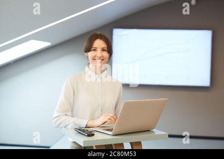 Waist up Porträt von lächelnden jungen Geschäftsfrau Blick auf Kamera, während posiert am Stehtisch mit Laptop in minimalem Innenraum mit Präsentation sc Stockfoto