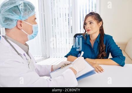 Ziemlich lächelnde junge Frau, die Empfehlungen des Arztes in medizinischer Maske und Gummihandschuhen hört Stockfoto