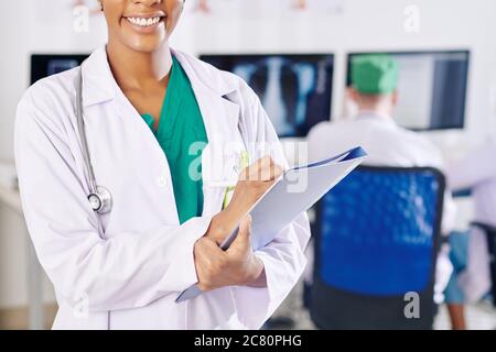 Zugeschnittenes Bild von schwarzer Ärztin mit schönen toothy lächelnd Fülldokument in ihren Händen Stockfoto