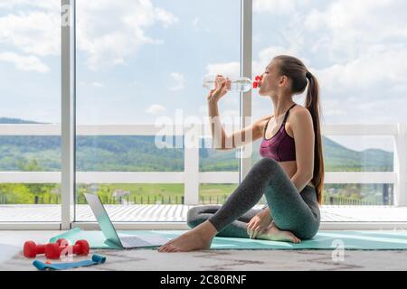 Eine sportliche Frau in Sportbekleidung sitzt mit Hanteln und Wasser auf dem Boden und benutzt zu Hause im Wohnzimmer einen Laptop. Sport und Stockfoto