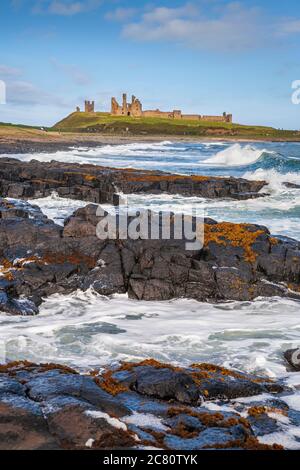 Wellen brechen auf den schwarzen Felsen von Dunstanburgh Castle, Northumberland, England Stockfoto