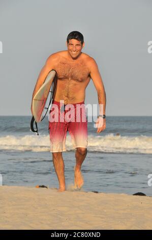 Porträt eines jungen, gutaussehenden Mannes in seinen Zwanzigern kommen Aus dem Meer heraus, ein Surfbrett unter seinem Arm tragend Und lächelnd