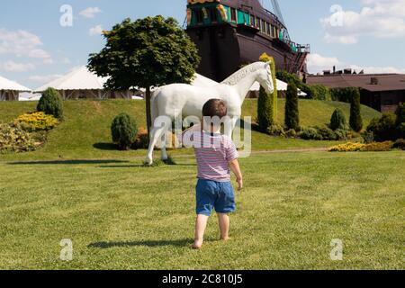 Ein 3-jähriger Junge im gestreiften T-Shirt barfuß auf dem Gras geht zu einem weißen Pferd. Sommer sonnige Kindheit Stockfoto