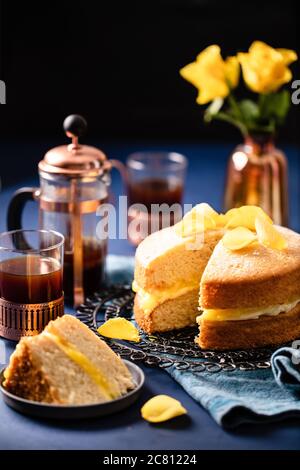 Dekadente Zitrone Biskuit mit gelben Rosenblättern mit frischem Kaffee auf tiefblauem Hintergrund serviert dekoriert Stockfoto