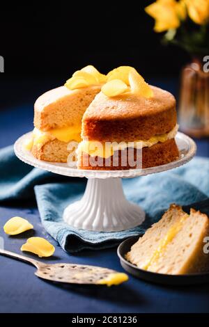 Dekadent veganer Zitronenkuchen präsentiert auf dekorativen weißen Kuchenständer, gekrönt mit gezuckerten gelben Rosenblättern. Kuchenscheibe auf Teller Stockfoto
