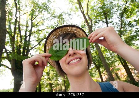 Fröhliche junge Frau trägt Blätter als Sonnenbrille und genießt die Natur an einem sonnigen Tag auf einer Waldlichtung Stockfoto