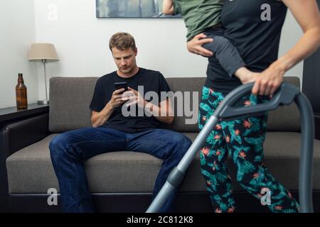 Fauler Mann sitzt auf der Couch und mit dem Telefon, während Frau Hausreinigung mit Kind auf ihrem Schoß Stockfoto