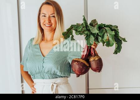 Junge Frau, die rote Beete mit grünen Blättern hält und lächelt an Kamera Stockfoto