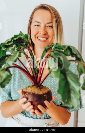 Junge Frau, die rote Beete mit grünen Blättern hält und lächelt an Kamera Stockfoto