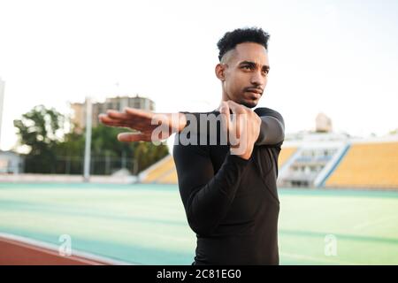 Junger selbstbewusster afroamerikanischer Sportler, der sich auf der Rennstrecke im Stadion nachdenklich dehnt Stockfoto