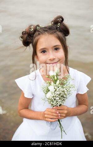 Ein Mädchen hält einen Strauß weißer Blumen auf einem verschwommenen Hintergrund.dunkles Haar, weiße Blumen in den Händen, weißes Kleid Stockfoto