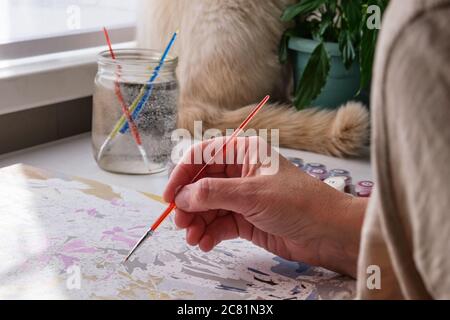 Die Hand der älteren Frau hält einen Pinsel. Sie malt mit Acrylfarben nach Zahlen auf Leinwand. Digitales Detox-Konzept Stockfoto