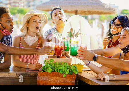 Multiethnische Freunde trinken Cocktails in einer Strandbar draußen in Sommertagen mit Gesichtsmaske auf vor Coronavirus geschützt werden - Happy People cheerin Stockfoto