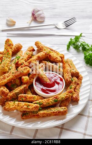 Nahaufnahme von knusprigen Zucchini-Sticks paniert mit Panko Paniermehl, Parmesan-Käse, Gewürze auf einem weißen Teller mit Ketchup auf einem Holztisch, vertikal Stockfoto