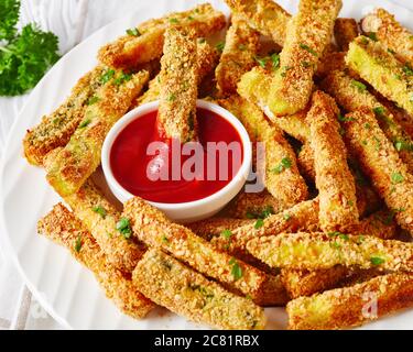 Nahaufnahme von knusprigen Zucchini-Sticks paniert mit Panko-Paniermehl, Parmesan-Käse, Gewürze auf einem weißen Teller mit Ketchup auf einem Holztisch, Makro Stockfoto