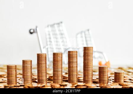 Goldene Münztürme mit gefallener Miniatur-Supermarkt-Warenkorb im Hintergrund, selektiver Fokus. Stockfoto