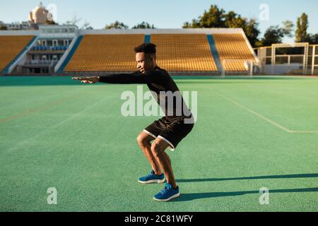 Seitenansicht des jungen afroamerikanischen Sportlers, der beim Training im Stadtstadion hockt Stockfoto