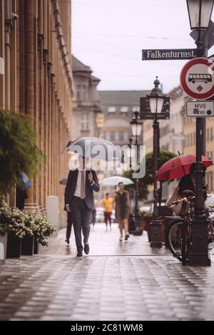 MÜNCHEN, 07. Jul 2020: Geschäftsmann in einem Anzug auf der Straße in der Stadt während des Regens. Mann mit Regenschirm in der Innenstadt von München. Stockfoto