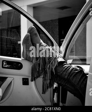 Ein Gewirr aus Wildleder fällt nach unten von einem Paar Denim gekleideten Lady Beine wanderte Ruhe gekreuzten Knöcheln an der Autotür tragen Mokassin Stil Stiefel Stockfoto