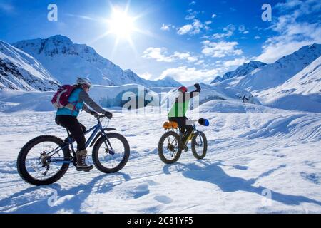 Zwei fette Frauen, die an einem sonnigen Wintertag vor dem Skookum Glacier, Chugach National Forest, Alaska, fahren, fist pumpend, während sie... Stockfoto