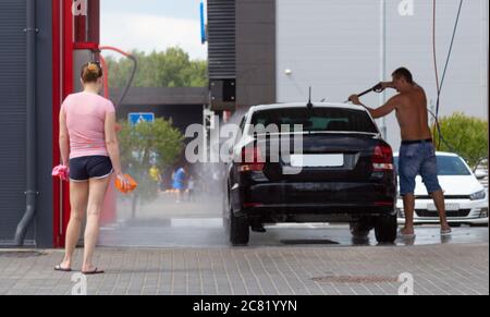 Ein Mann wäscht sein Auto mit einem Schlauch unter dem Druck von Wasser. Die Frau steht in der Nähe. Autowäsche Stockfoto