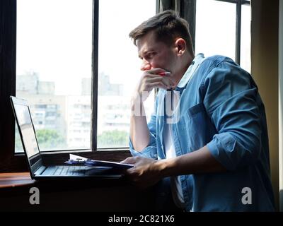 Junger attraktiver Mann am Fenster, der am Laptop arbeitet. Freiberufliche und Remote-Arbeit. Stockfoto