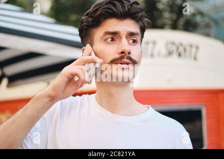 Porträt eines jungen, nachdenklichen Schnurrbart-Mannes im T-Shirt, der auf dem Handy auf der Straße spricht Stockfoto