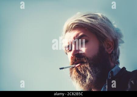 Serious bärtiger Mann rauchen eine Zigarette am Strand. Bärtige Hipster am Himmel. Nahaufnahme im Hochformat. Stockfoto