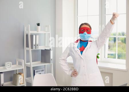 Der Arzt in einer Schutzmaske hob im Büro der Klinik die Hand in einem Superheldenanzug hoch Stockfoto