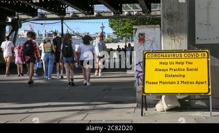 London, Großbritannien. Juli 2020. Gruppen von Menschen gehen an einem sozialen distanzierenden Erinnerungszeichen in der South Bank vorbei. An vielen öffentlichen Orten in London gibt es Schilder, die sich auf 2 Meter Entfernung halten, aber die Regeln werden nicht immer eingehalten, da viele Menschen die Entspannung der Absperrung und Geselligkeit bei warmem Wetter genießen. Kredit: Imageplotter/Alamy Live Nachrichten Stockfoto