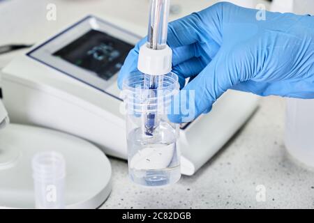 Frau in einem Gummihandschuh mit einer Glaselektrode zur Messung des pH-Wertes der Lösung mit einem pH-Messgerät. Labor für analytische oder elektrochemische Untersuchungen. Stockfoto