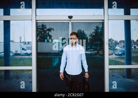 Ein junger kaukasischer Geschäftsmann mit Sonnenbrille steht vor dem Flughafen mit einer schwarzen Aktentasche in der Hand Stockfoto