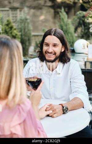 Junge attraktive Brünette bärtigen Mann im Hemd mit Glas weinfreudig Blick auf Freundin auf Datum im Café im Freien Stockfoto