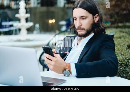 Junger selbstbewusster bärtiger Geschäftsmann mit einem Glas Wein, der im Restaurant im Freien mit einem Handy nachdenklich telefoniert Stockfoto