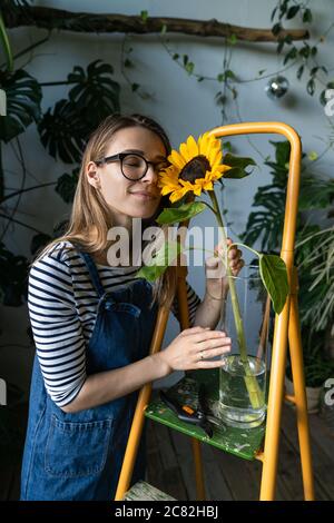Floristin Frau von tropischen Pflanzen umgeben genießt eine schöne Sonnenblume in Vase, in der Nähe der orangefarbenen Trittleiter stehen. Stockfoto