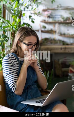 Frau Gärtnerin in Brille tragen blaue Jeans Overalls, sitzen auf Stuhl im Gewächshaus, mit Laptop nach der Arbeit, kommuniziert im Internet mit Kunden su Stockfoto