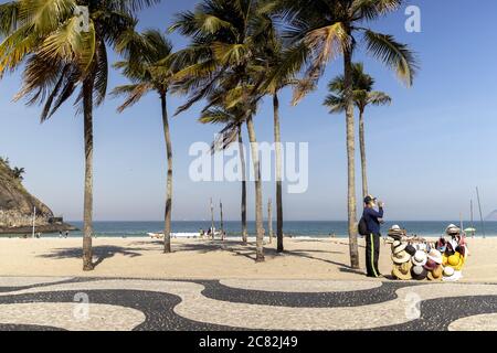 RIO DE JANEIRO, BRASILIEN - Jul 06, 2020: Leme Boulevard mit Straßenverkäufer trägt Gesichtsmaske Verkauf Hüte pausieren unter Palmen, um Wasser zu trinken o Stockfoto