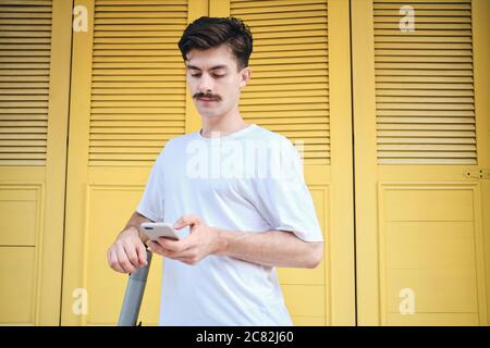 Junger Schnurrbart Mann in weißem T-Shirt nachdenklich mit Handy mit gelbem Hintergrund auf der Straße Stockfoto