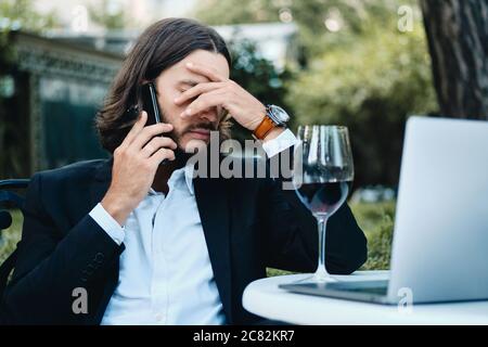 Junge verärgert Geschäftsmann mit einem Glas Wein und Laptop frustrierend auf Handy im Restaurant im Freien reden Stockfoto