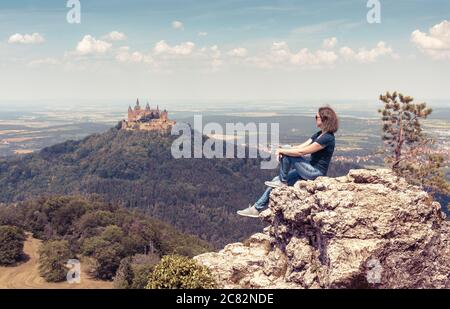 Junge Frau sitzt auf einem Felsen in der Nähe von Schloss Hohenzollern. Dieses gotische Schloss ist Wahrzeichen in der Nähe von Stuttgart. Hübsches Mädchen touristisch reist in moun Stockfoto