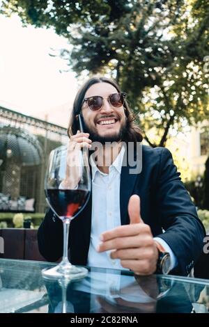 Junge gutaussehende lächelnde Brünette bärtigen Mann freudig auf Handy reden, während mit einem Glas Wein im Restaurant im Freien ruhen Stockfoto