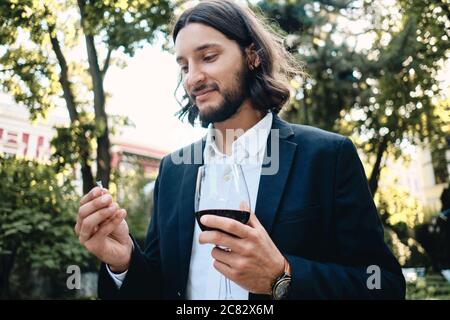 Junger, hübscher bärtiger Mann mit einem Glas Wein, der im Restaurant im Freien einen Trauring hält Stockfoto